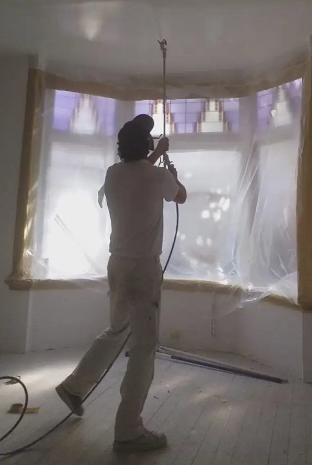 Latexspuiter spuit een plafond met een airless spuit op verlengstuk; erker en glas-in-lood zijn afgeschermd met folie en tape